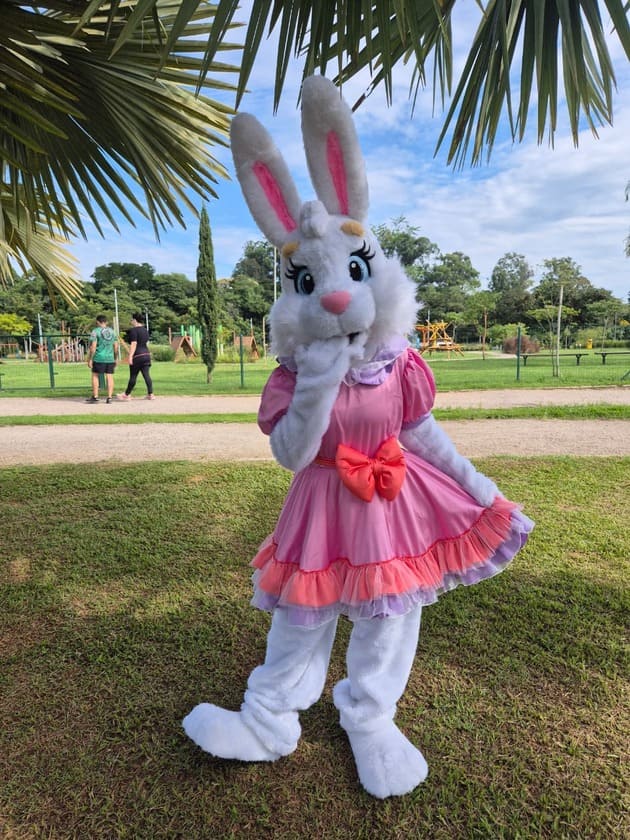 coelha branca com roupa rosa com salmão colocando a mão na boca, com outra mão estica a saia, enquanto passa duas pessoas atras.