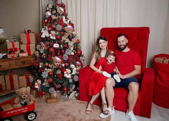 Família sentada em poltrona dupla vermelha para Papai Noel ao lado de árvore de Natal decorada