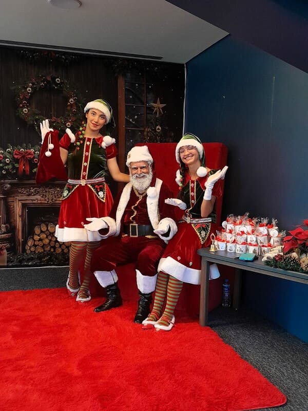 Papai Noel acompanhado por duas Noeletes em poltrona dupla vermelha durante evento de Natal