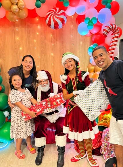 Papai Noel e duas Noeletes com família durante entrega de presentes em evento de Natal