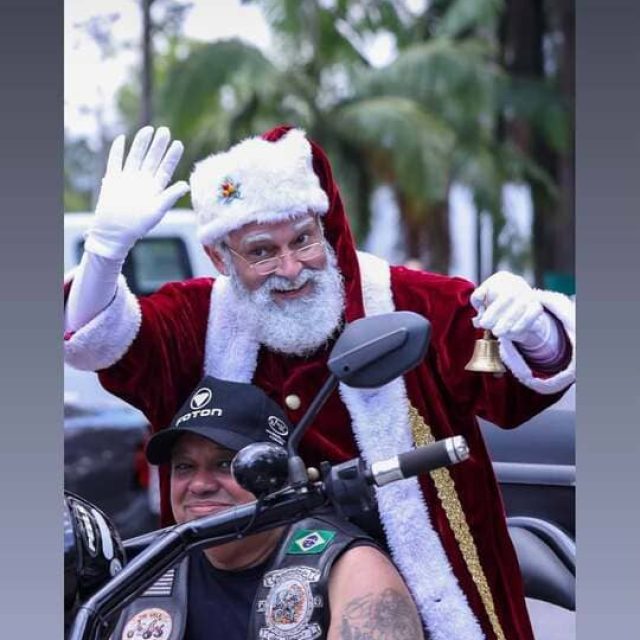 Papai Noel com barba natural acenando e segurando um sino durante evento ao ar livre
